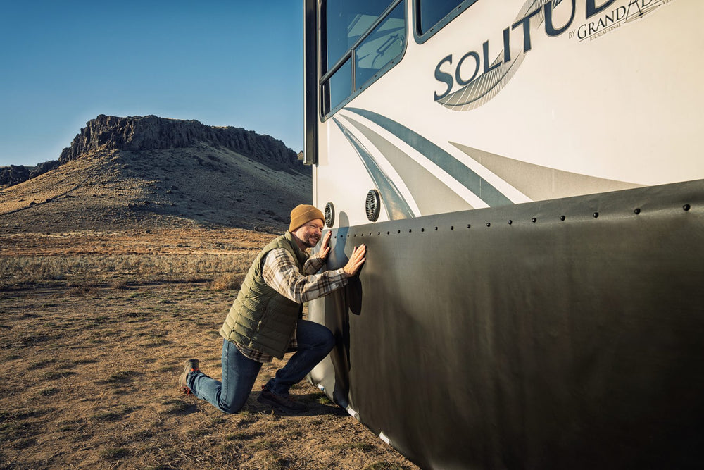 Man kneeling beside an RV, pressing black EZ Snap skirting into place along its side
