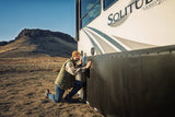 Man kneeling beside an RV, pressing black EZ Snap skirting into place along its side